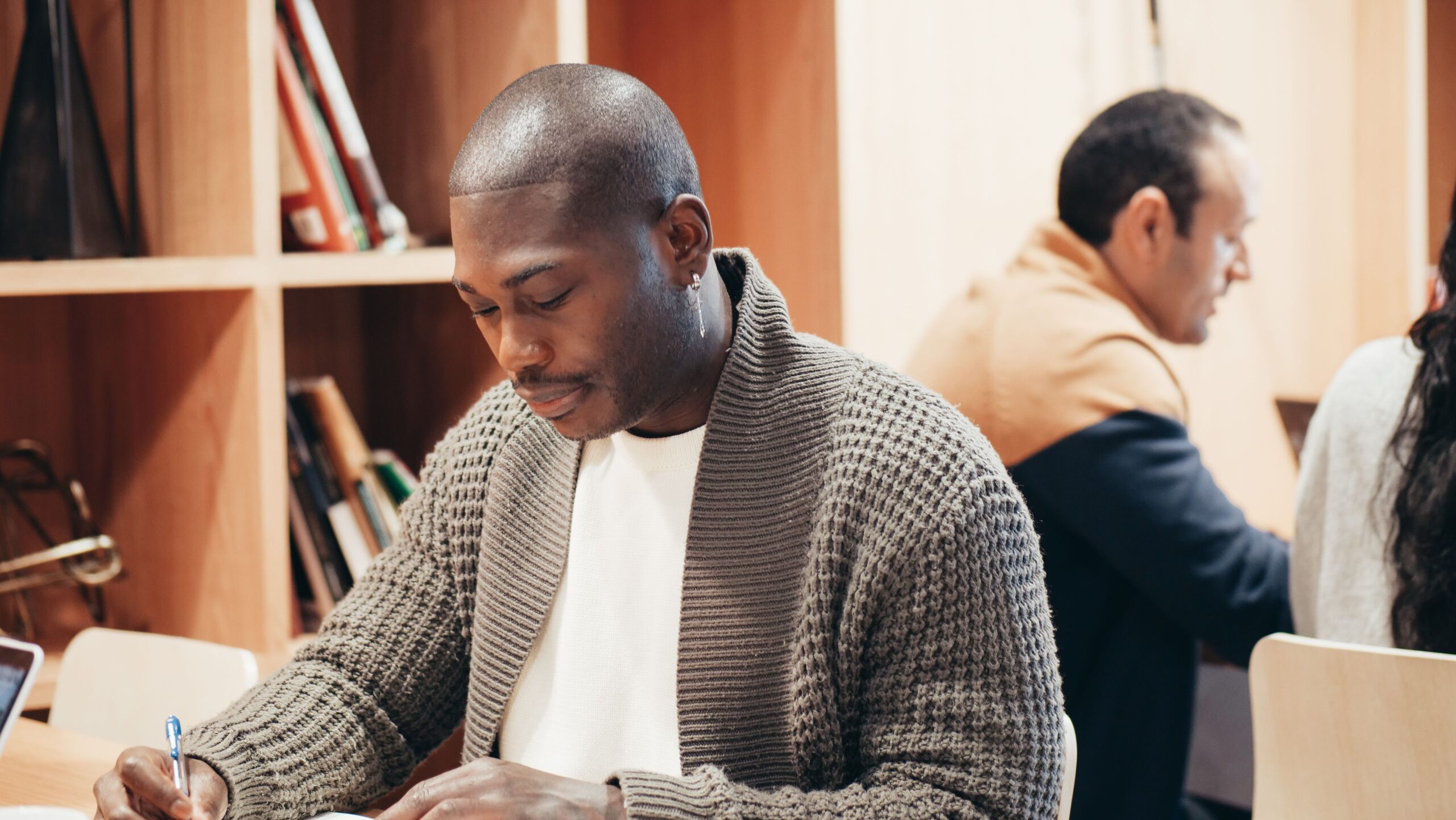 An adult man writing at a desk in a cozy office environment with bookshelves.